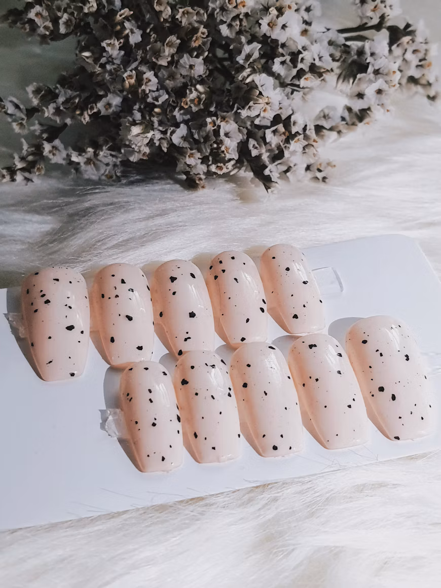 Ten cream press-on nails with a black speckled design on a display card.